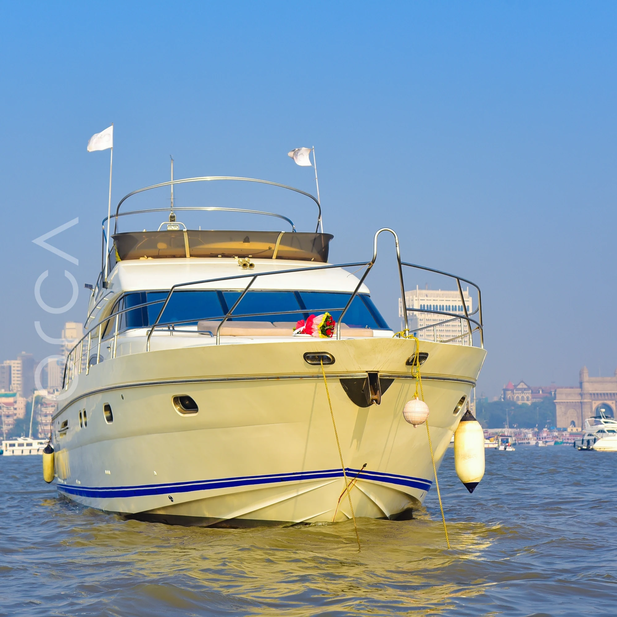 The Princess 61 motor yacht at anchor in Mumbai harbour, bow three-quarter view with the Gateway of India behind, available to rent from Orca Sail.