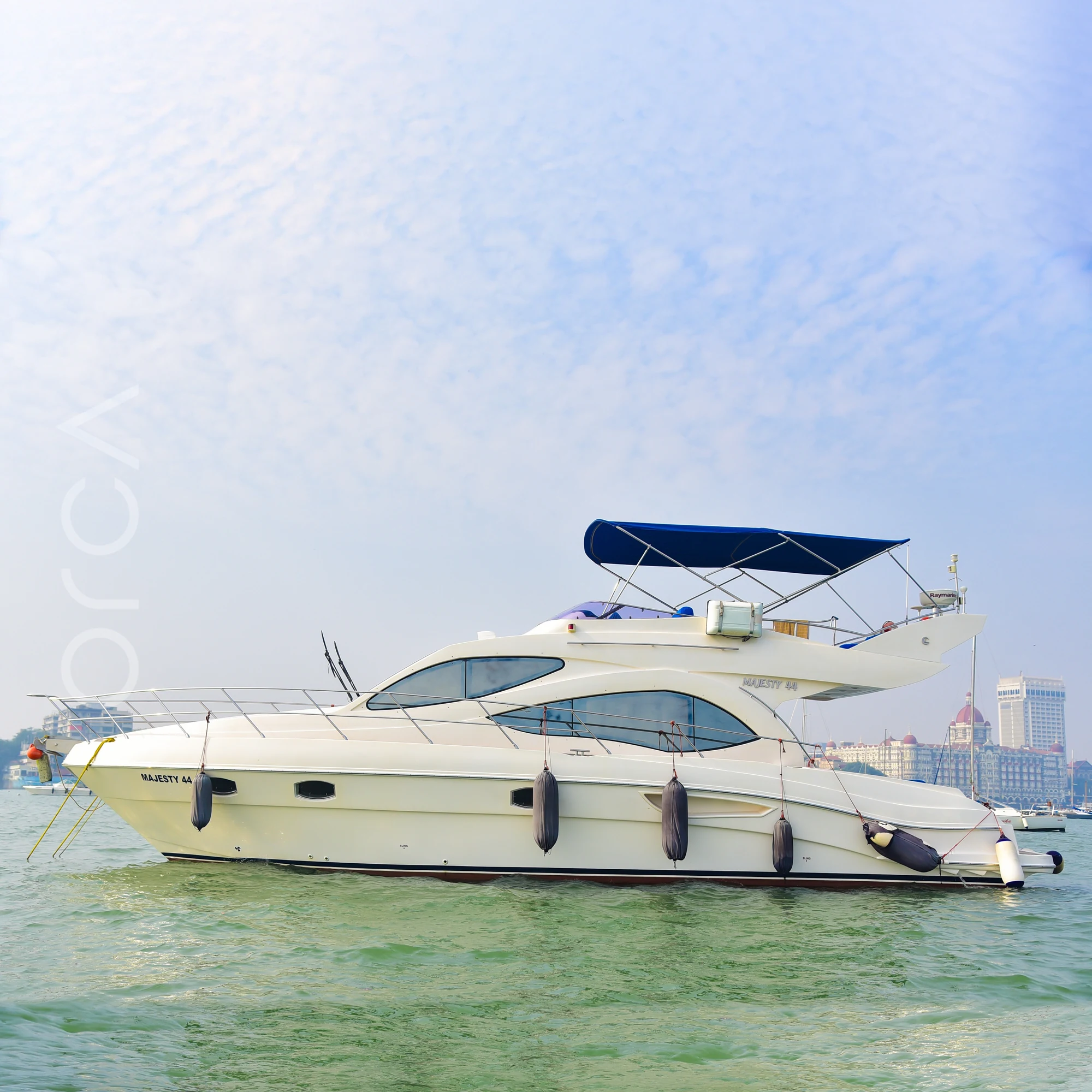 The full length of a Majesty 44 motor yacht seen side-on in Mumbai harbour, with the Taj Mahal Palace behind, available to rent from Orca Sail.